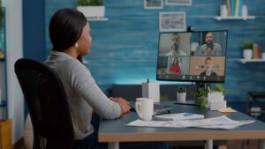 Remote Work, a photo of a woman with her back to the camera looking at a screen divided into four with an individual person displayed in each sector. The desk is very tidy with one plant and in the background past the computer monitor is a blue all with a few shelves of trinkets on it.